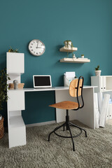Interior of stylish home office with laptop, workplace and clock hanging on blue wall