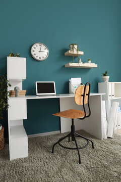 Interior of stylish home office with laptop, workplace and clock hanging on blue wall