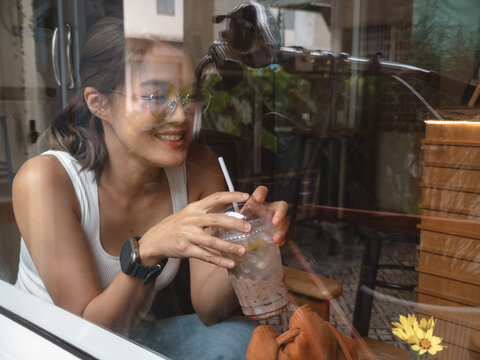 Happy Young Asian Woman with Glasses Sips Iced Coffee in Cafe, Seen Through Window