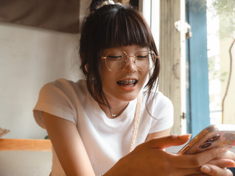 Happy young Asian woman with glasses and braces smiling while using her smartphone indoors.