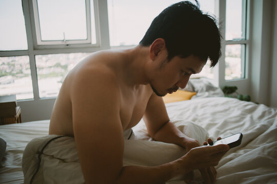 Shirtless Asian man focused on his mobile phone while relaxing in bed at home.
