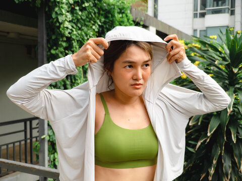 Focused young woman in sportswear putting on a light jacket in a green outdoor environment.
