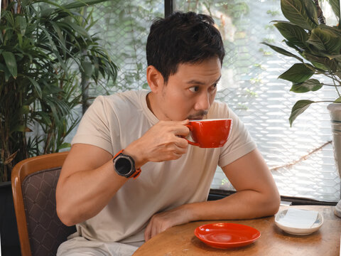 Young Asian Man Enjoying a Hot Beverage in a Cozy Cafe Setting, Looking Away Thoughtfully