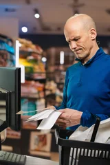 Bald caucasian man frowning at checkout while holding receipts and basket, close to register and scanner, expression shows disbelief at total cost and hesitation at payment moment. © Denys Kurbatov