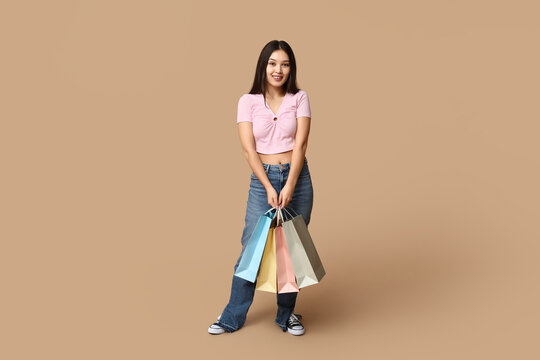 Fashionable young Asian woman in stylish jeans with shopping bags on brown background