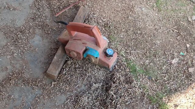 Commercial stock video showing an orange portable electric wood planer power tool sitting on a dirty wooden board in a workshop.