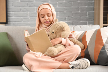Fototapeta premium Little Muslim girl in hijab with teddy bear reading book at home