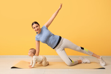 Fototapeta premium Beautiful young sporty woman with her little baby practicing yoga near yellow wall