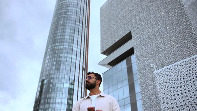 Bearded man tapping smartphone while standing in urban plaza beside glass skyscraper and patterned concrete architecture, with man, beard, smartphone, phone detail