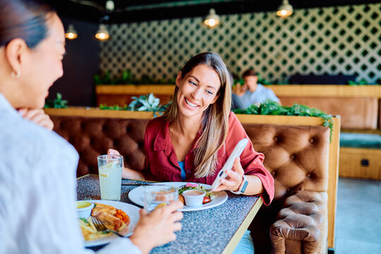 Two happy women friends laughing and looking at a smartphone while enjoying lunch together at a modern restaurant