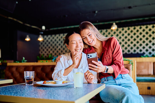 Two cheerful women having lunch at a restaurant, sharing a moment of joy while looking at a smartphone screen