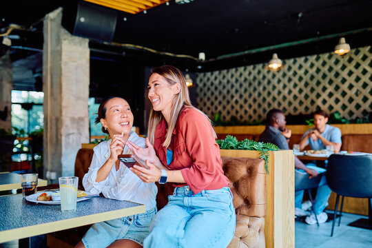 Two cheerful women having a good time, talking and laughing over drinks and food in a modern restaurant interior
