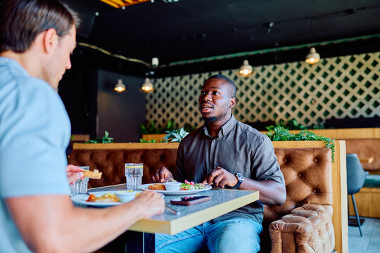Two young men, one African American, eating and talking at a restaurant table. Represents casual friendship and dining out