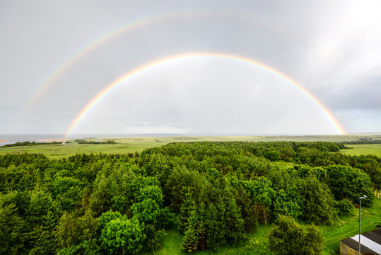 Spectacular double rainbow arching over lush green forests, golf fields and coastal countryside near the North Sea after rain under dramatic cloudy skies