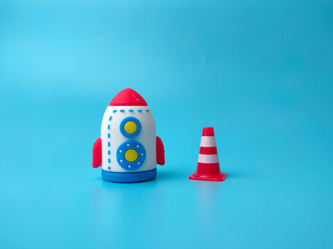 A toy rocket placed beside a small red and white traffic cone on a blue background