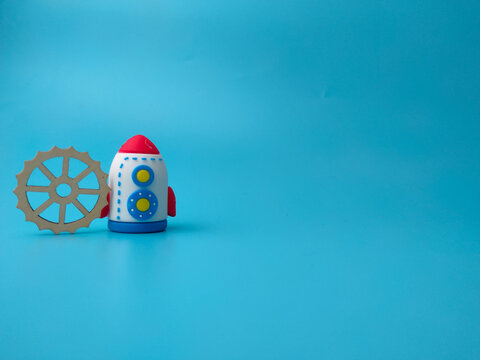 A toy rocket standing next to a wooden gear cogwheel against a solid blue background