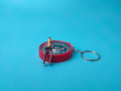A miniature figurine of a businessman sitting on a red pocket compass with a keychain ring over a blue background