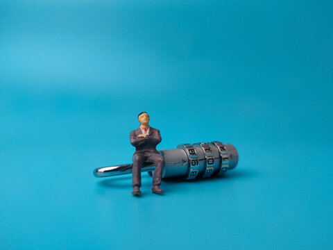 A miniature businessman sitting on top of a silver combination padlock against a plain blue background