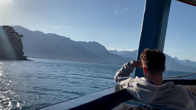A young teenage boy is sailing on a ship. View of the Taurus mountain range. The Mediterranean Sea in Kemer, Turkey. View from a ship in the open sea. Seascape. Blue water. 4k