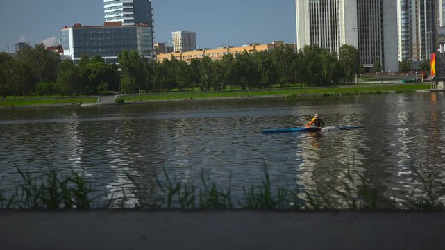 Two kayakers paddling across reflective city river, leaving wakes and ripples that break tall building reflections during sunny daylight from low waterfront viewpoint
