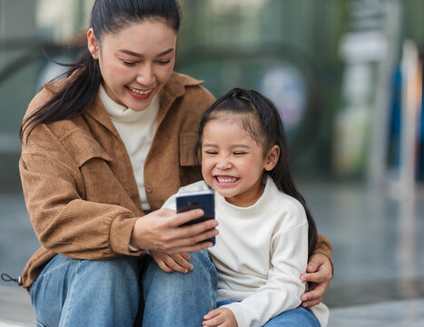 happy mother and daughter sitting down and looking at smartphone in city
