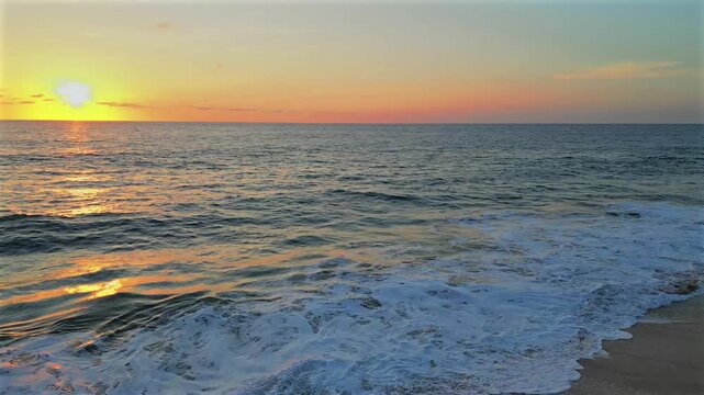 Drone flies low over Secret Rocks Beach below Todos Santos, moving from the sand to the open ocean at sunset, passing just above a wave as it breaks and rolls beneath.