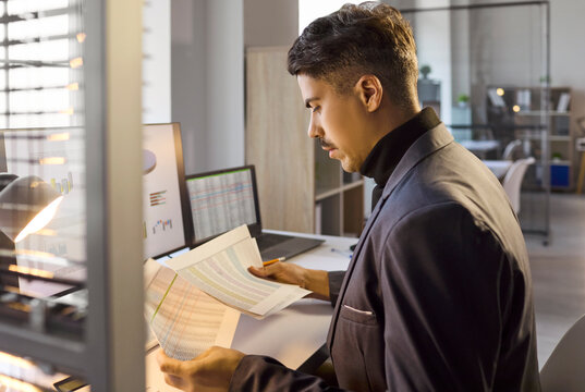 Businessman analyst in office reviews accounting documents. He is a manager auditing charts beside a monitor and laptop, checking audit figures. Clear concept of finance and business analysis.