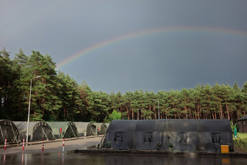 Obraz premium A rainbow arcs over rows of green army tents beside a pine forest after a rain shower at a military camp.