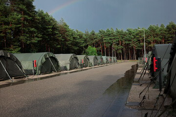 Obraz premium A rainbow rises over rows of green army tents and a pine forest after a summer rain shower at a military camp.