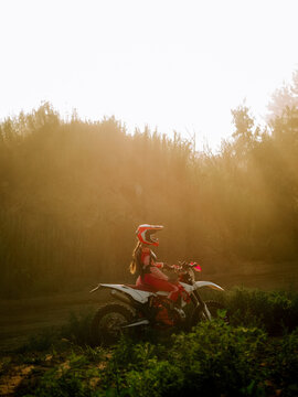 Motocross rider sitting on a white dirt bike during golden hour in a forest clearing at sunset