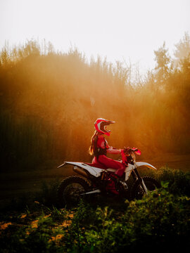 Motocross rider in red gear sits on a dirt bike during a bright and golden sunset in a forest area