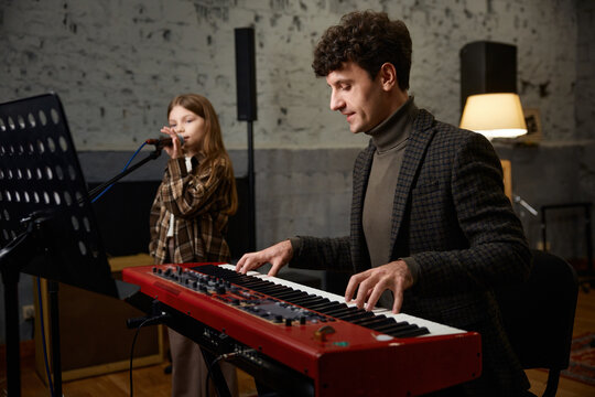 Caucasian coach accompanies girl vocalist at mic during focused studio rehearsal