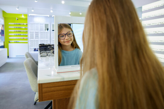 Charming girl selecting glasses in mirror with cheerful expression