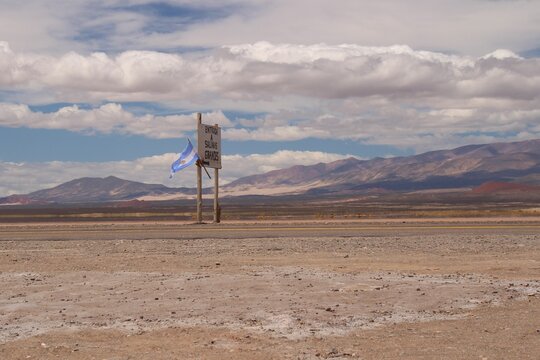 Entrance to the Salinas Grandes salt flats in northwestern Argentina, marked by a rustic roadside sign and an Argentine flag fluttering in the wind