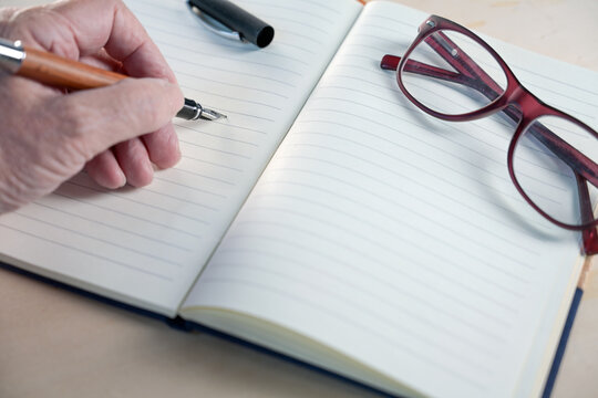 Older hand with a fountain pen writing in a notebook with blank lined paper, glasses laying on the side, concept image for handwriting, diary, notes, and creative writing, copy space,
