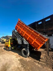 Fototapeta premium Orange Dump Truck Unloading Soil at an Active Construction Site Under a Clear Blue Sky