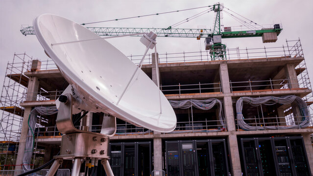 Large white satellite dish antenna positioned in front of a modern construction site. Building frame features server racks and a tower crane. Data center infrastructure concept for tech marketing.