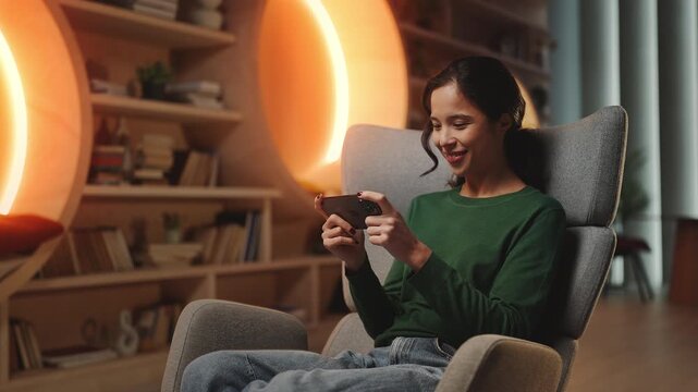 Cheerful young Korean woman playing video game on phone in home study, hobby