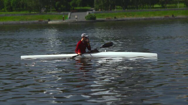 Adult paddler in white racing kayak pausing for hydration before resuming powerful stroke on calm urban river beneath bright midday sunlight and reflections near park banks
