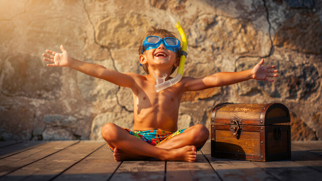 Happy boy in snorkel mask sits with open arms by a treasure chest. Child celebrates imaginary underwater adventure in sunlight. Fun childhood exploration concept for travel and summer ads.