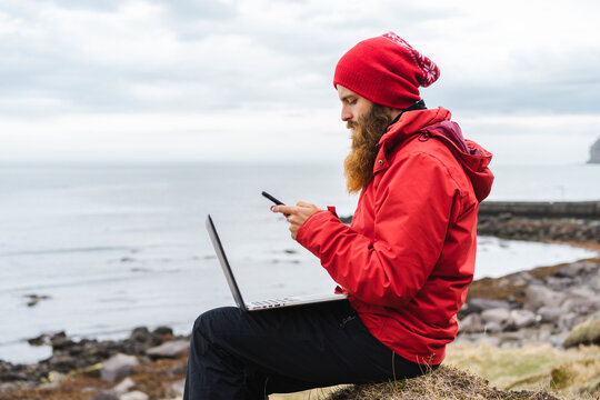 Iceland, North of Iceland, man with laptop sitting near the sea using cell phone