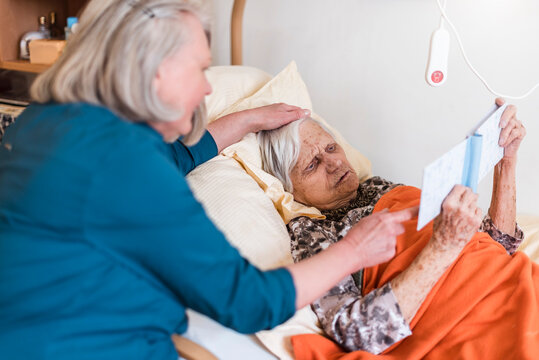 Woman taking care of old woman lying in bed reading book