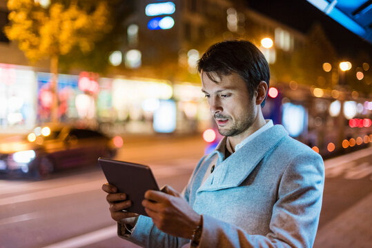 Businessman with digital tablet standing at a bus stop at night