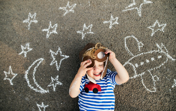 Smiling toddler with pilot hat and goggles lying on asphalt painted with airplane, moon and stars