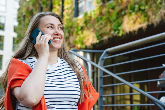Smiling woman talking on smartphone outdoors near green architecture