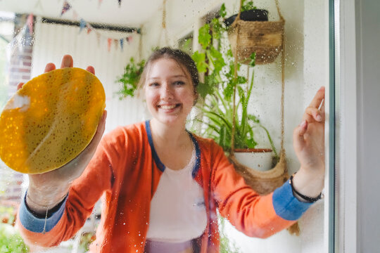 Smiling teenage girl cleaning door with sponge on balcony
