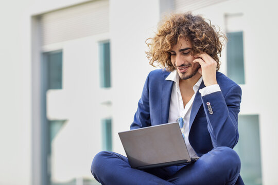 Smiling young businessman with curly hair wearing blue suit sitting on bench outdoors looking at laptop