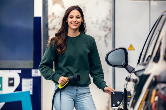 Happy young woman standing with electric plug at vehicle charging station