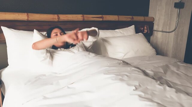 Young woman wakes up in a comfortable bed, stretches her arms, and relaxes under a white duvet in a cozy bedroom. She turn off air condition while enjoying peaceful morning at home and doze off again