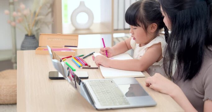 Side view of Asian single mother tutoring little daughter through drawing task with pink pencil near laptop, phone, notebook, and color set, supporting creativity, attention, and caring home routine.